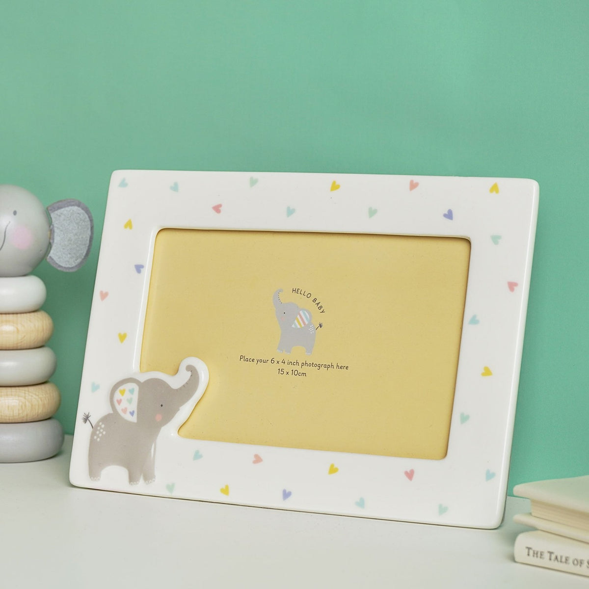 White ceramic photo frame with pastel heart border and raised grey elephant at the corner, holding a yellow 6x4 inch placeholder, displayed against a green background.