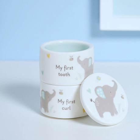Stacked round white ceramic keepsake pots labelled "My first tooth" and "My first curl", decorated with grey elephants, bees and hearts, with lid resting beside them.