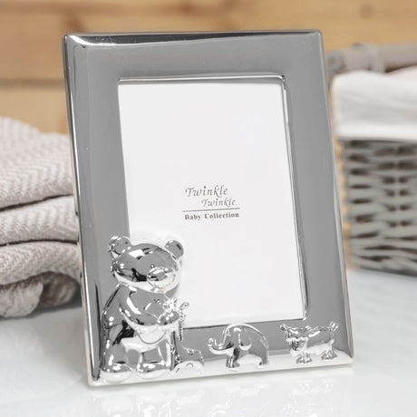 Silverplated rectangular photo frame for a 5 x 3.5in portrait photo, with a raised teddy bear, elephant and lamb along the bottom edge, shown on a table near towels and a basket.