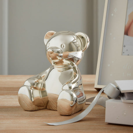 Sitting Teddy Bear Silverplated Metal Money Box with polished silver finish, legs stretched out, shown on a wooden surface beside a photo frame and gift box.