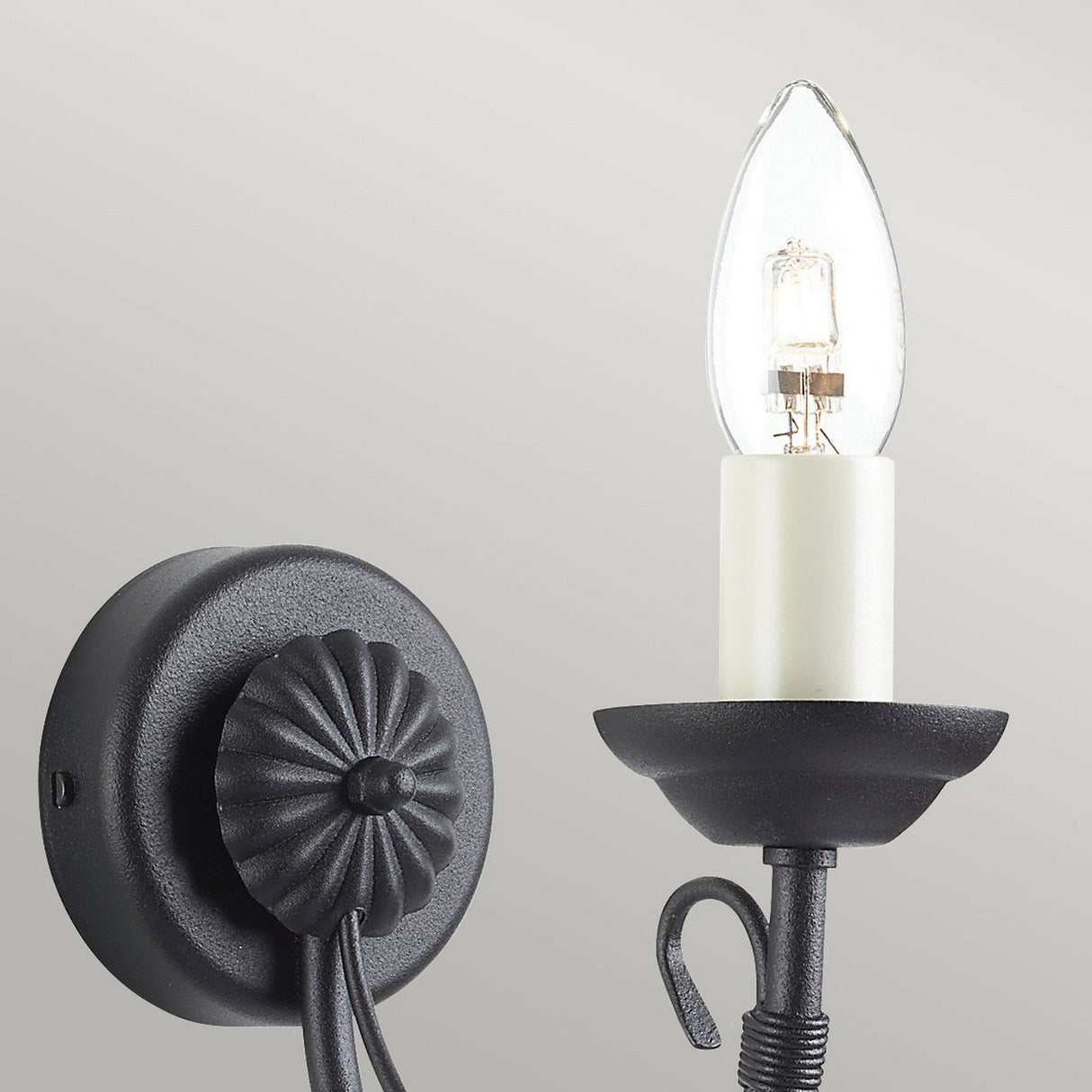 The Chartwell 1 Light Wall Light in black features a vintage-style candelabra design with a decorative circular base and a single exposed bulb resembling a candle flame. Its arm is adorned with a small curved detail, set against a simple, light gray wall.