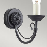 The Chartwell 1 Light Wall Light - Black showcases a black metal wall sconce with a decorative circular base and an upward-curving arm that supports a single bulb in a white holder. Its traditional candelabra style is simple and elegant, making it ideal for classic or rustic decor.