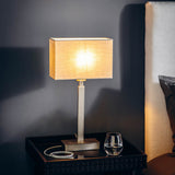 Chartstead rectangular 27cm tapered lamp shade in grey fabric with rolled edge, shown lit on a table lamp beside a bed in a bedroom setting.
