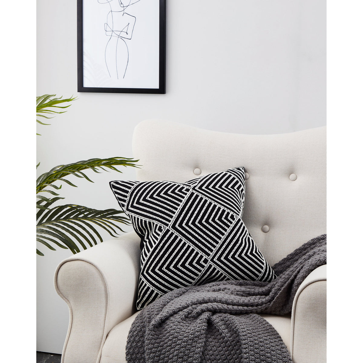 Chevery Cushion in black and white with a textured chevron and diamond pattern, styled on a light upholstered armchair beside a grey throw and framed line art.