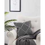 Chevery Cushion in black and white with a textured chevron and diamond pattern, styled on a light upholstered armchair beside a grey throw and framed line art.