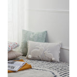 Chevery rectangular cushion in natural and white with sherpa-fleece rabbit design and pompom tails, shown on a patterned rug beside a green patterned cushion.