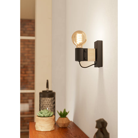 Cirbette Wall Light in black steel and light brown wood, with an exposed E27 bulb and black braided flex, shown mounted on a white wall above a wooden sideboard.