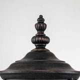 Close-up view of the top of a black, ornamental outdoor fixture featuring a decorative finial. The Cleveland-1 Outdoor Medium Wall Lantern showcases clear seeded glass in a vintage, weathered bronze finish. The background is plain white.