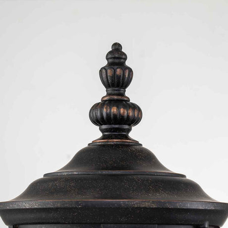 Close-up view of the top of a black, ornamental outdoor fixture featuring a decorative finial. The Cleveland-1 Outdoor Medium Wall Lantern showcases clear seeded glass in a vintage, weathered bronze finish. The background is plain white.