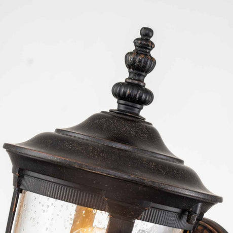 A close-up of the Cleveland-1 Outdoor Small Wall Lantern reveals a decorative black metal top with clear seeded glass panels. The weathered bronze finish, complete with rustic details, highlights its vintage charm.