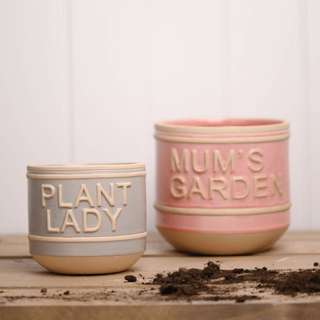 Set of 2 round ceramic planters with raised beige lettering, a grey "Plant Lady" pot and a pink "Mum's Garden" pot, both with beige rim bands and bases, shown on wood with scattered soil.