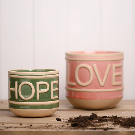 Set of 2 gloss ceramic planters with embossed lettering, a green HOPE pot and a larger pink LOVE pot, both with sandy beige raised bands and curved feet, shown on a wooden surface with scattered soil.