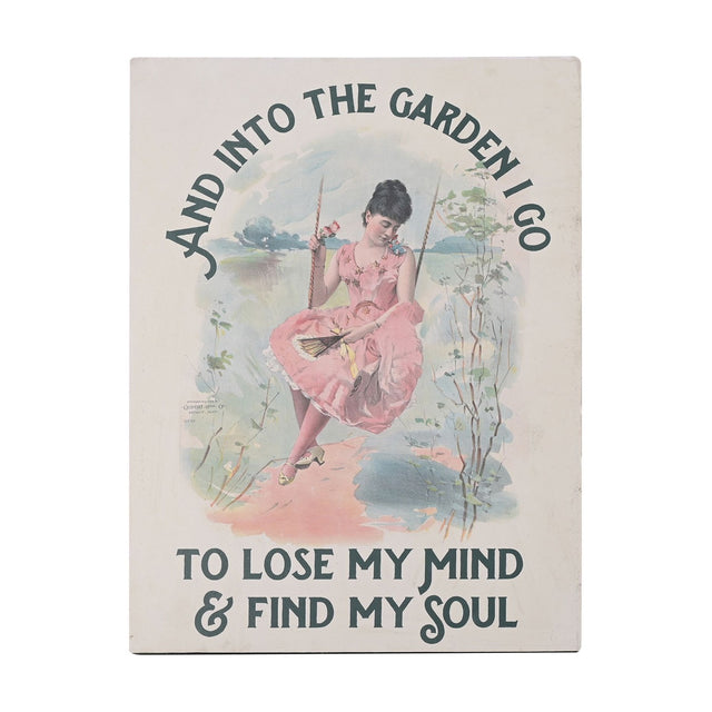 Cream metal wall plaque, 40cm, with arched dark slate text reading "And into the garden I go to lose my mind & find my soul" around an illustration of a woman in a pink dress on a swing.