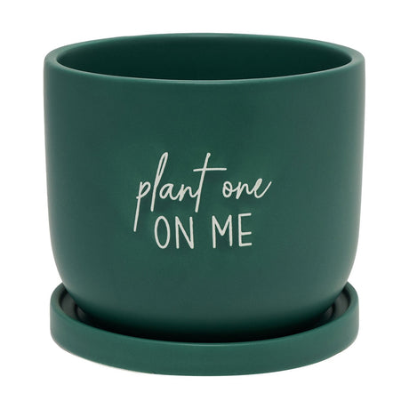 Matt green ceramic planter with curved rounded sides and "plant one ON ME" in white script lettering, set on a matching green saucer against a white background.