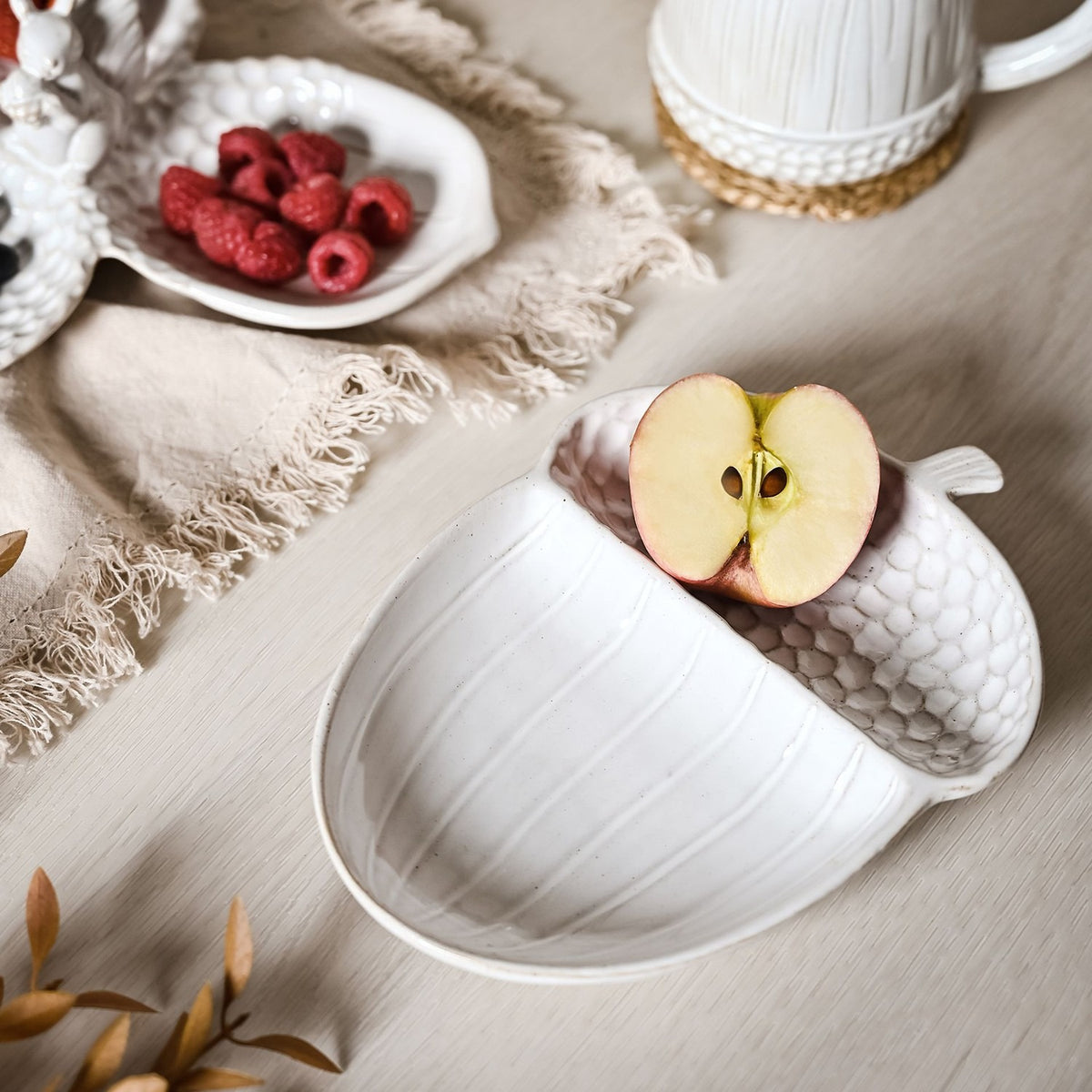 White glazed ceramic acorn-shaped serving plate with a pebbled textured cap and ribbed body, shown holding a halved apple beside raspberries on a linen-draped table.
