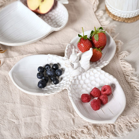 White glazed ceramic serving tray with three acorn-shaped dishes joined by a squirrel figure, holding blueberries, strawberries and raspberries on a fringed cloth.
