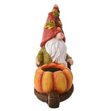 Rear view of the ceramic gonk garden ornament in a red leaf-patterned hat, white beard, and olive jacket, pushing an orange pumpkin wheelbarrow planter on a plain white background.