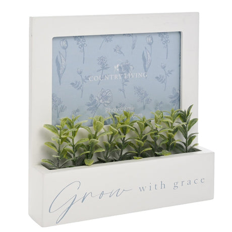White MDF wooden photo frame with landscape opening and base planter trough holding faux green leafy stems, text reads "Grow with grace".