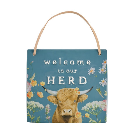 Ceramic wall plaque in teal with raised white "welcome to our HERD" text, a Highland cow illustration, and painted meadow flowers, shown with a leather hanging strap on a white background.