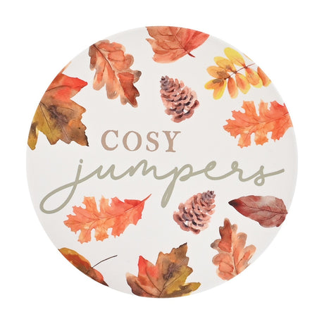 Round white ceramic coaster featuring painted autumn leaves and pine cones around "COSY jumpers" text in taupe and sage grey, shown on a plain background.