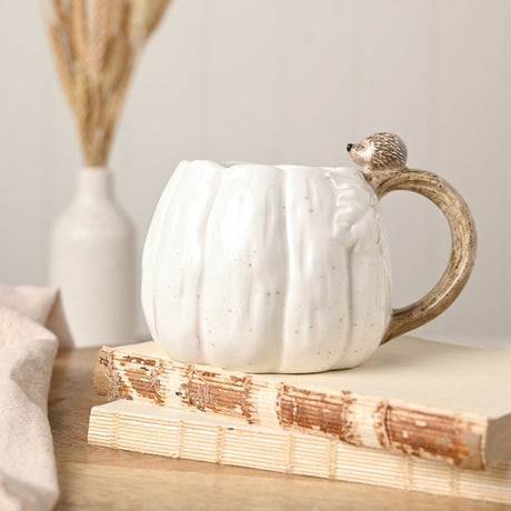 White ceramic pumpkin mug, 450ml, with ribbed body, leaf detail, and a branch-shaped handle topped with a small hedgehog figure, shown on books with dried flowers.