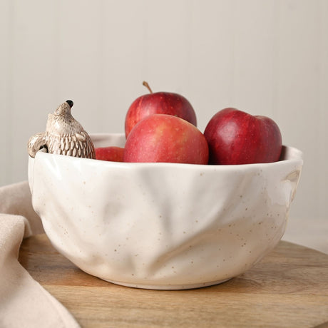 Round white speckled ceramic serving bowl with dimpled, uneven sides and a small hedgehog figure perched on the rim, filled with red apples on a wooden surface.