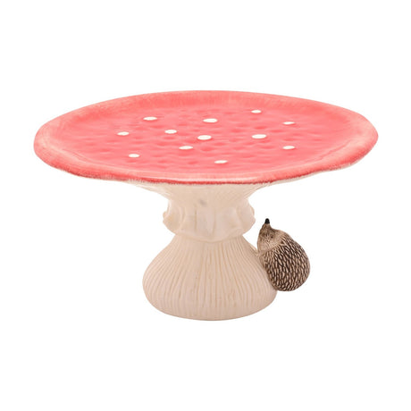 Ceramic cake stand shaped like a red toadstool with white spots on a round dimpled cap, ribbed cream stalk, and a small brown hedgehog figure on the side, shown on a plain white background.