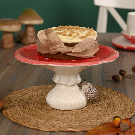 Ceramic footed cake stand styled as a red, white-spotted toadstool with a ribbed cream stalk and a hedgehog figure, shown holding a cake on a table.