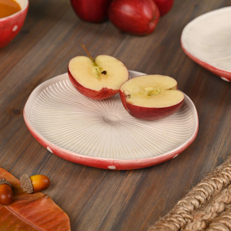 Round ceramic side plate with ribbed toadstool-gill texture in white and a red rim, shown on a wood table with sliced apple halves on top.