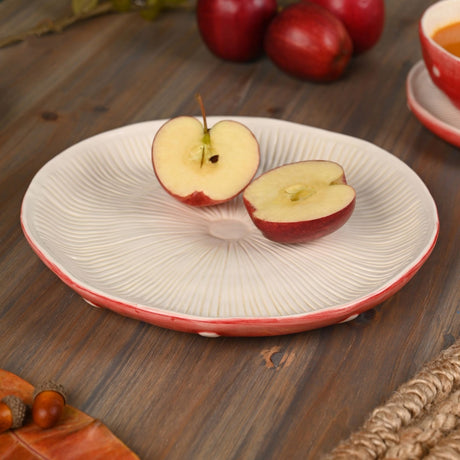 Round ceramic dinner plate with textured cream centre and red, white-spotted rim, shown on a wooden table with sliced apples.