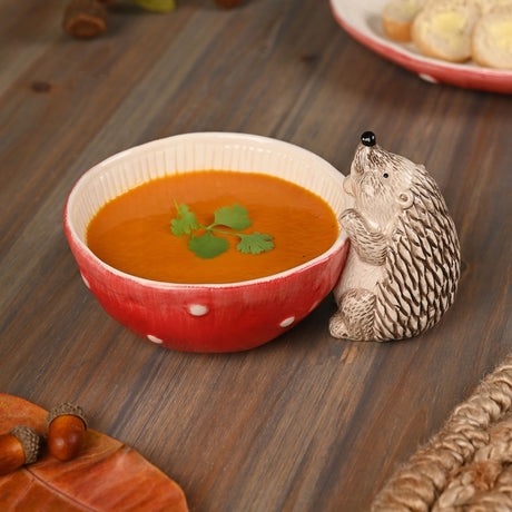 Round Toadstool & Hedgehog Serving Bowl, 450ml, in red with white polka dots and a cream ribbed interior, shown filled with orange soup beside its cream hedgehog figure on a wooden table.