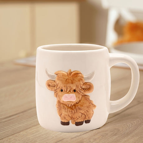 Cream stoneware barrel mug with a raised highland cow design, featuring a ginger-brown fringe, pink nose and pale horns, shown on a wooden table in a kitchen setting.