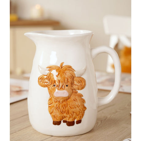 White stoneware jug with a raised, hand-painted Highland cow in ginger-brown, featuring a pink nose and pale horns, wide pouring lip, and loop handle, shown on a wooden table in a home setting.