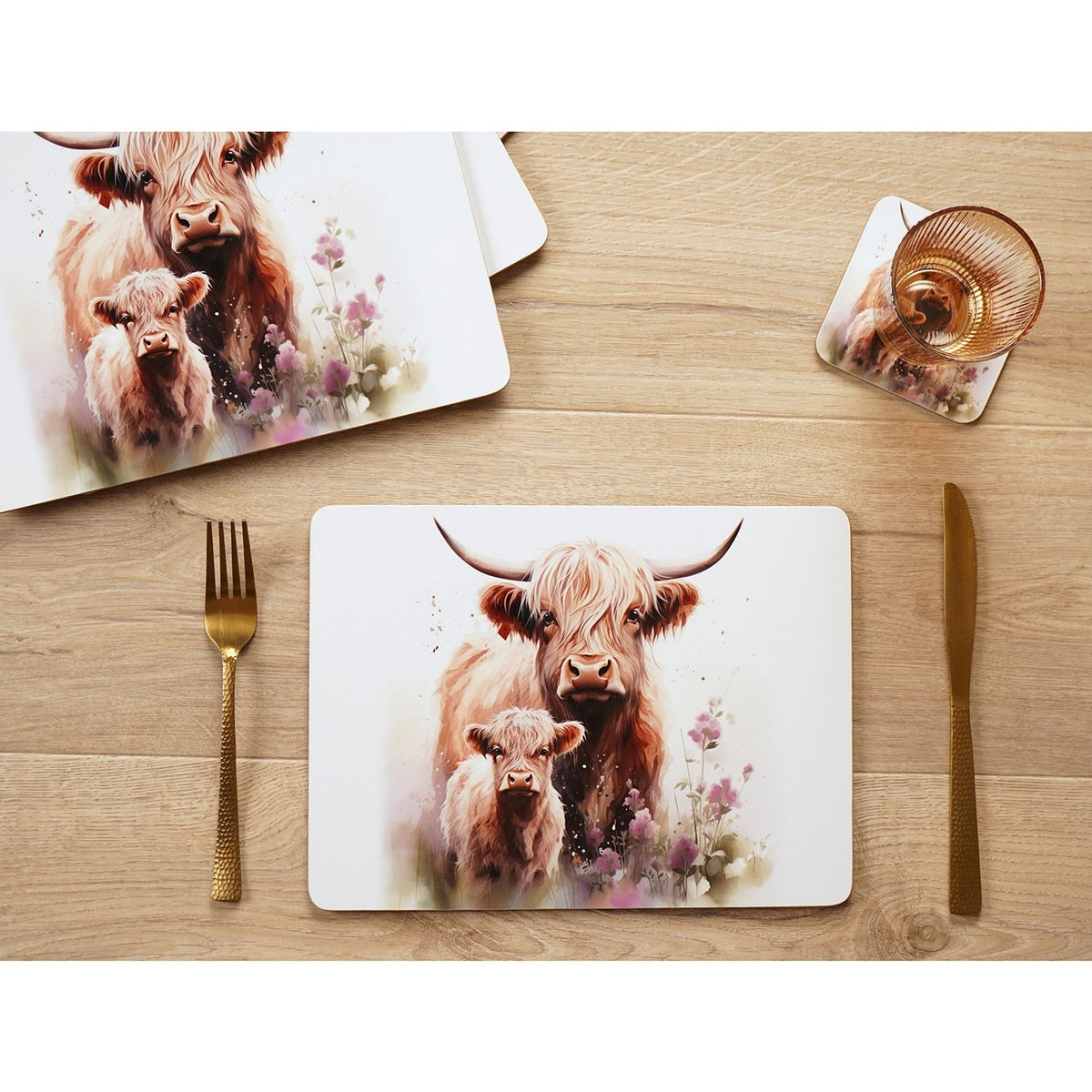 Rectangular placemat printed with a watercolour Highland cow and calf among pink wildflowers, set on a wooden table with cutlery and a matching coaster visible nearby.