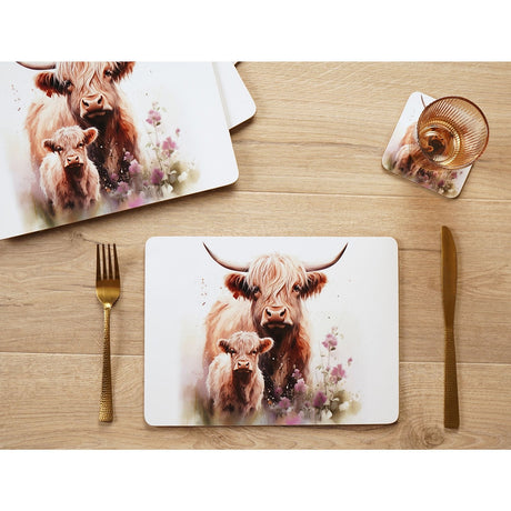 Rectangular placemat printed with a watercolour Highland cow and calf among pink wildflowers, set on a wooden table with cutlery and a matching coaster visible nearby.