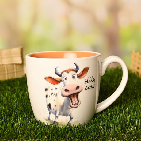 White earthenware mug with orange interior featuring a cartoon cow and "silly cow" text, shown outdoors on artificial grass.