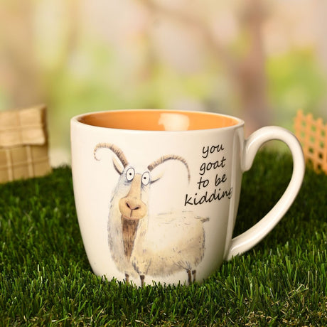 White earthenware mug with an illustrated goat and "you goat to be kidding" text, orange glazed interior, shown outdoors on grass.