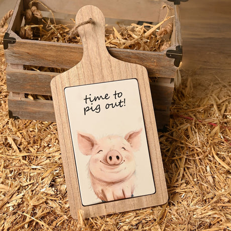 Paddle-shaped earthenware serving board with light brown wood-look frame and cream ceramic tile, featuring a sketched smiling pig and "time to pig out" script, shown on straw beside a wooden crate.