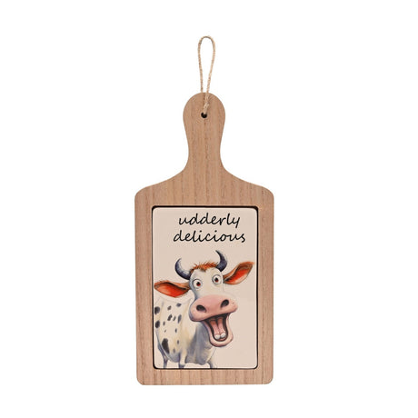 Wooden paddle-shaped serving board with rounded handle and jute hanging cord, set with a ceramic tile featuring a grinning cartoon cow and "udderly delicious" text, on a white background.