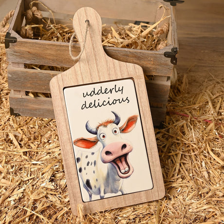 Wooden paddle-shaped serving board with ceramic tile insert featuring a smiling cow illustration and "udderly delicious" text, shown leaning against a straw-filled crate on hay.