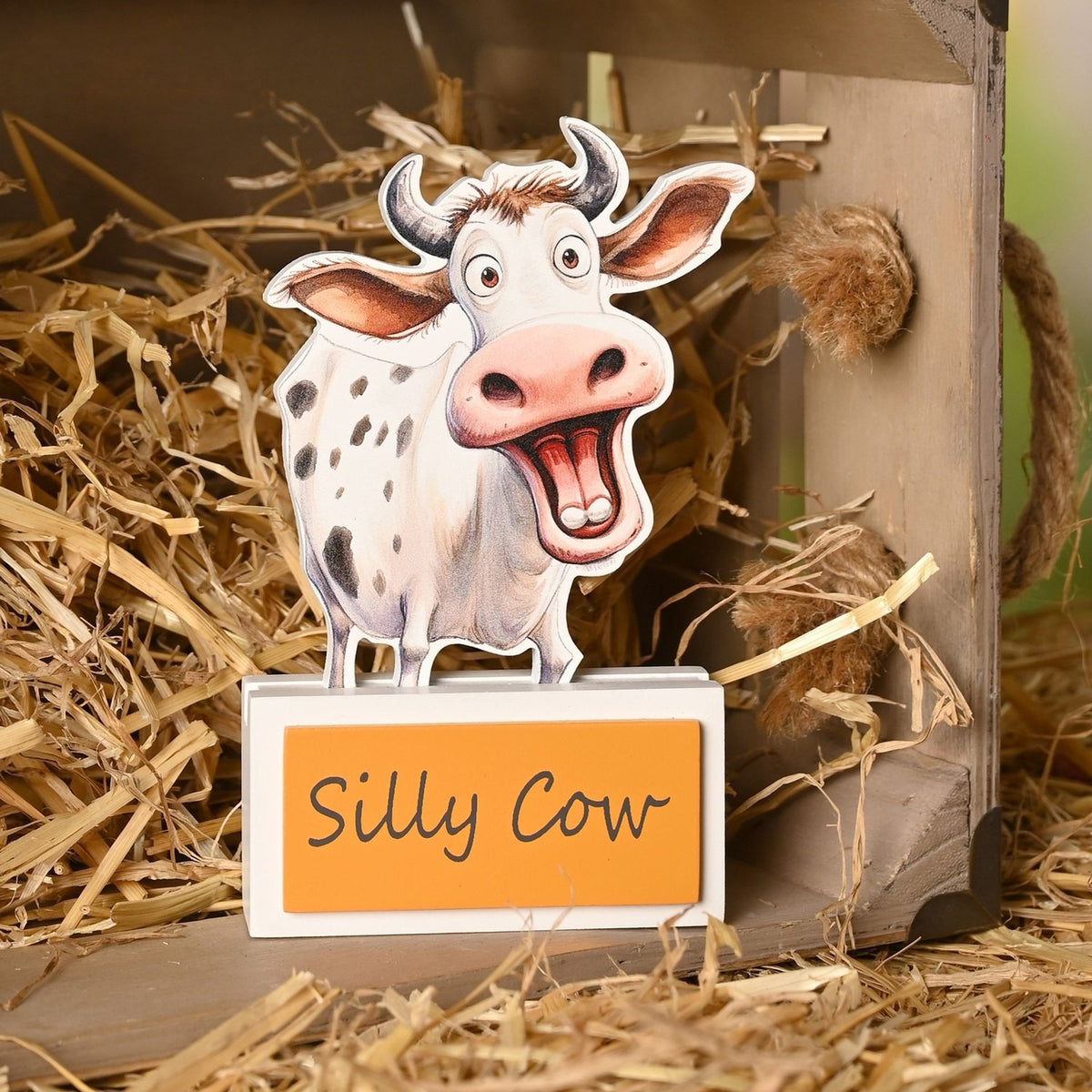 Wooden cut-out plaque of a cartoon cow with spotted coat and open-mouthed grin, standing on a white base with orange "Silly Cow" panel, set among straw.