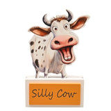 Wooden standing plaque cut to a cartoon cow's outline with spotted white coat, pink nose and grey horns, on a white base with an orange "Silly Cow" panel, shown against a plain white background.