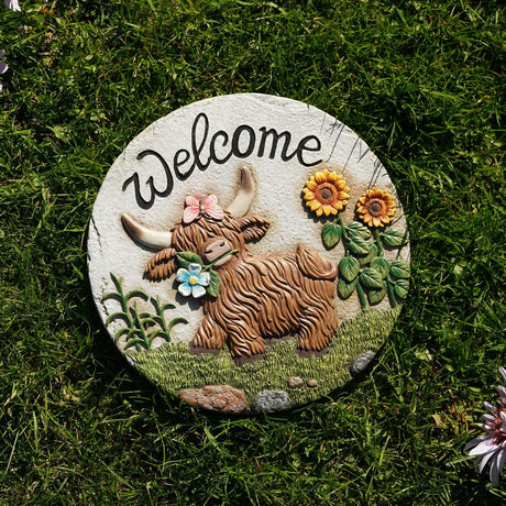 Round resin stepping stone with "Welcome" text and a raised Highland cow design, pink butterfly, blue flower and sunflowers, in grey and brown finish, set in grass.