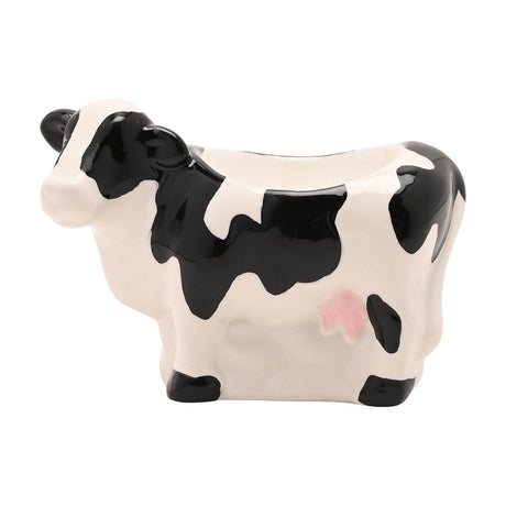 Standing Cow Earthenware Egg Cup in glossy white with black patches, black hooves and ears, and a pink udder, egg-holding hollow on its back, shown on a plain white background.