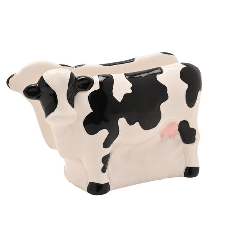 White and black ceramic napkin holder shaped like a standing spotted cow with black ears, nose, hooves and patches, pink udder, and a slot along its back for napkins.