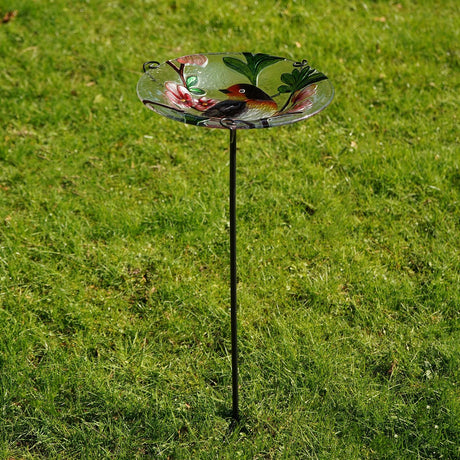 Glass bird feeder on a slim metal stake, with a round dish featuring a painted robin, pink blossom, and green leaves, set on green lawn.