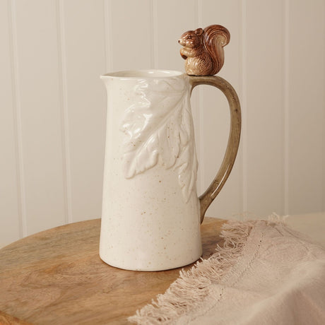 Cream ceramic jug with embossed oak leaf design and a brown squirrel figure holding an acorn perched on the curved handle, shown on a wooden board with a linen cloth.