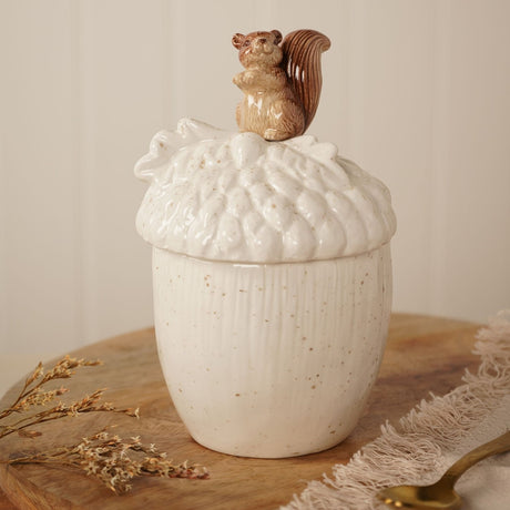 Cream ceramic acorn-shaped storage jar with lid, featuring a textured scaled cap, oak leaf detail, and a brown squirrel figure on top, displayed on a wooden board.