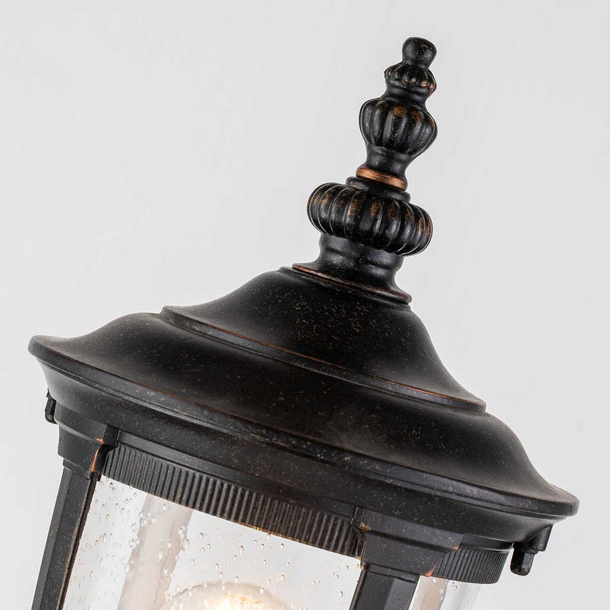 Introducing the Cleveland Small Pillar Lantern in Weathered Bronze—a stunning vintage-style outdoor light fixture. This lantern features a decorative finial and clear, textured glass panels. It beautifully complements a plain, light-colored background, making it an ideal choice for elegant outdoor lighting.