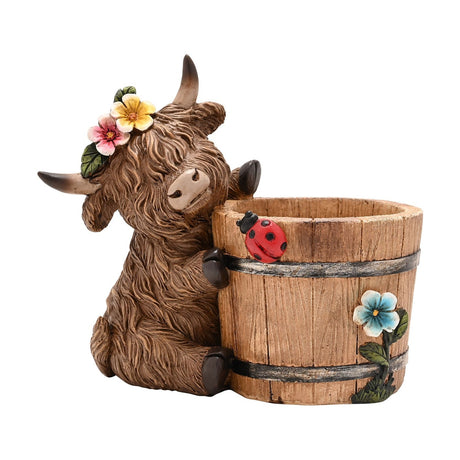 Highland Cow and Barrel Resin Planter in brown, a sitting cow with floral crown hugging a wooden-effect barrel decorated with a ladybird and blue flower, on a white background.